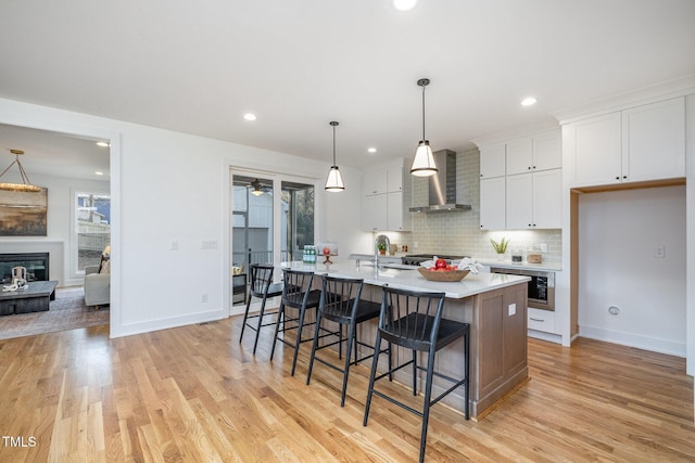 kitchen featuring wall chimney range hood, pendant lighting, white cabinets, light hardwood / wood-style floors, and an island with sink