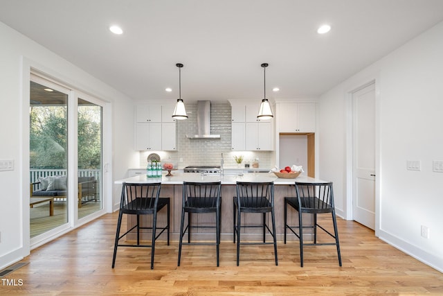 kitchen featuring wall chimney range hood, light hardwood / wood-style floors, a breakfast bar, white cabinets, and a center island with sink