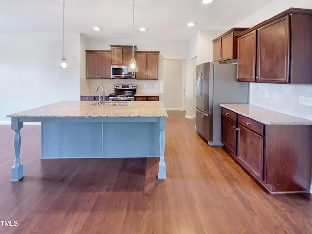 kitchen featuring hardwood / wood-style floors, a kitchen island with sink, decorative light fixtures, light stone counters, and stainless steel appliances