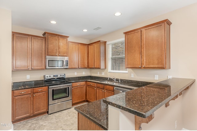 kitchen featuring a peninsula, a sink, appliances with stainless steel finishes, dark stone counters, and brown cabinetry