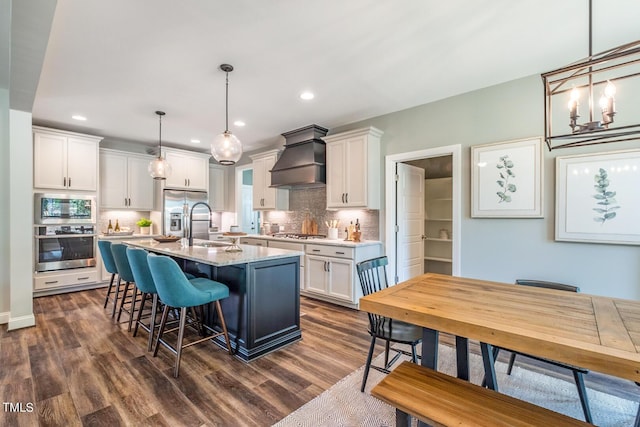 kitchen featuring dark hardwood / wood-style flooring, custom exhaust hood, stainless steel appliances, white cabinetry, and an island with sink