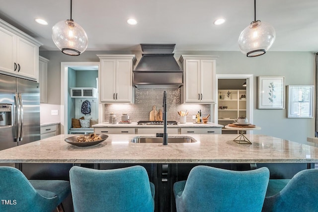 kitchen featuring stainless steel fridge, decorative light fixtures, custom range hood, and a center island with sink