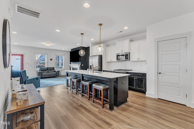 kitchen featuring light wood-type flooring, stainless steel appliances, pendant lighting, white cabinets, and an island with sink