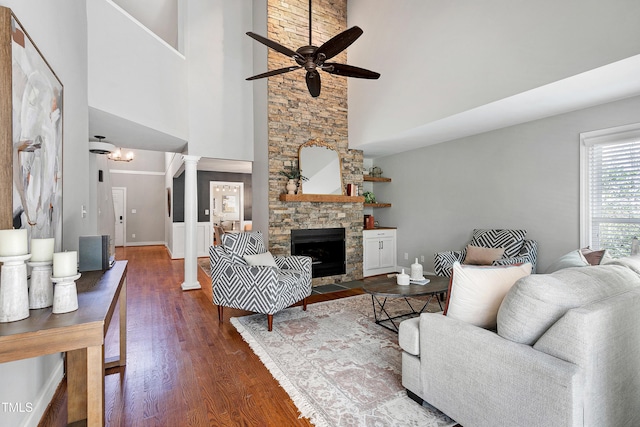 living area with dark wood-style flooring, a towering ceiling, a ceiling fan, a stone fireplace, and ornate columns