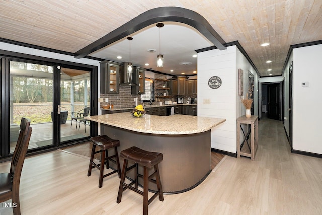 kitchen with dark brown cabinetry, wall chimney exhaust hood, light stone counters, a kitchen island, and light wood-type flooring