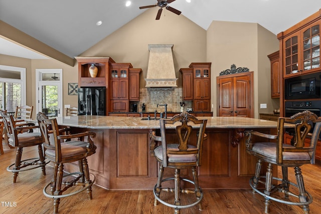 kitchen featuring a kitchen breakfast bar, hardwood / wood-style floors, an island with sink, black appliances, and custom range hood