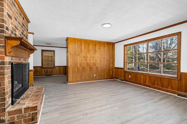 unfurnished living room with a brick fireplace, a textured ceiling, ornamental molding, wooden walls, and light hardwood / wood-style floors