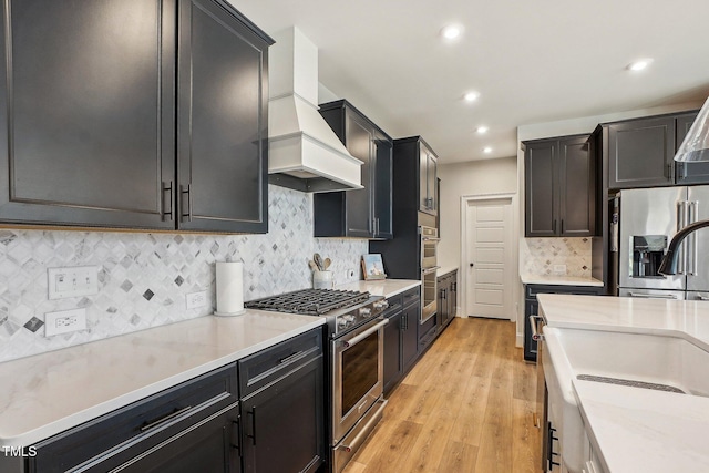 kitchen featuring light stone countertops, appliances with stainless steel finishes, light hardwood / wood-style floors, premium range hood, and decorative backsplash