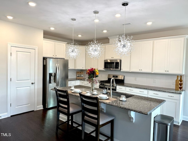 kitchen with appliances with stainless steel finishes, dark wood-type flooring, pendant lighting, white cabinetry, and an island with sink