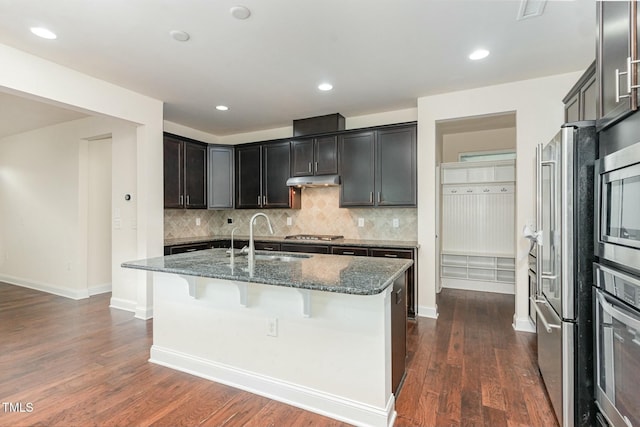 kitchen with dark wood-type flooring, sink, dark stone countertops, appliances with stainless steel finishes, and an island with sink