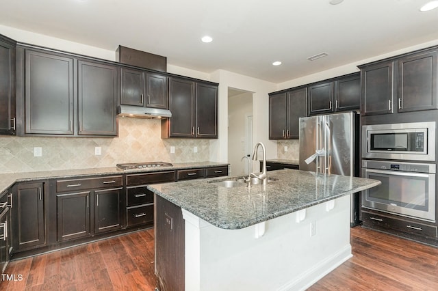 kitchen with dark wood-type flooring, sink, dark stone counters, stainless steel appliances, and a kitchen island with sink