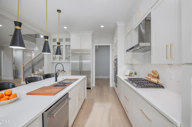 kitchen featuring appliances with stainless steel finishes, wall chimney range hood, white cabinetry, sink, and hanging light fixtures