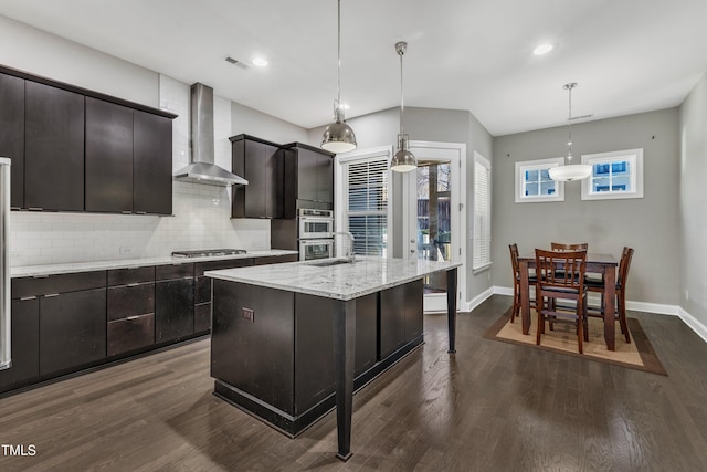 kitchen featuring an island with sink, backsplash, hanging light fixtures, stainless steel appliances, and wall chimney exhaust hood