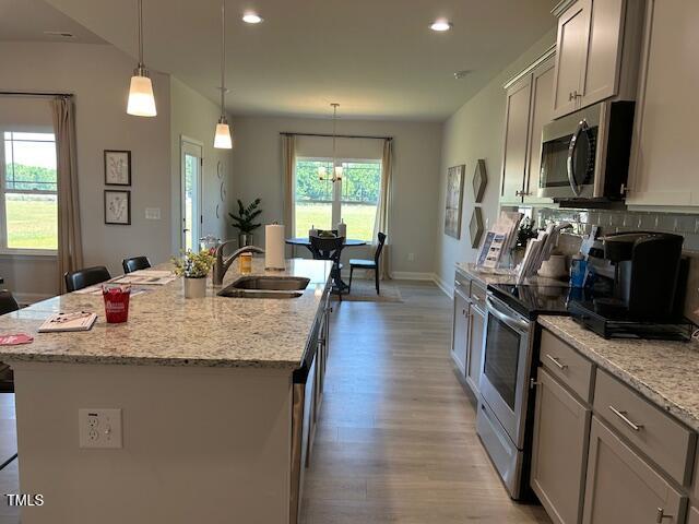 kitchen with a center island with sink, sink, hanging light fixtures, appliances with stainless steel finishes, and light stone counters