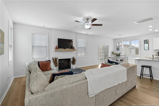 living area with baseboards, visible vents, a glass covered fireplace, light wood-style floors, and ceiling fan with notable chandelier