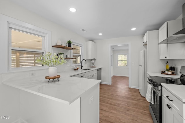 kitchen with wall chimney range hood, sink, white cabinetry, stainless steel appliances, and kitchen peninsula