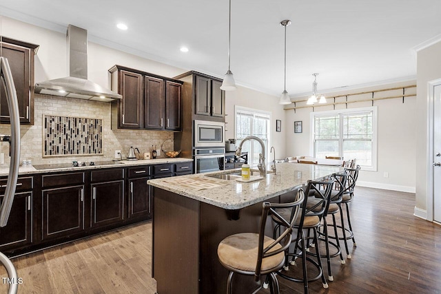 kitchen with appliances with stainless steel finishes, ornamental molding, a kitchen island with sink, sink, and wall chimney range hood