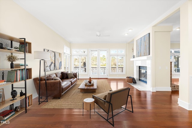 living area with ceiling fan, dark wood-type flooring, a fireplace with flush hearth, baseboards, and crown molding