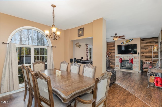 dining space featuring a tile fireplace, ceiling fan with notable chandelier, dark wood-type flooring, and wooden walls