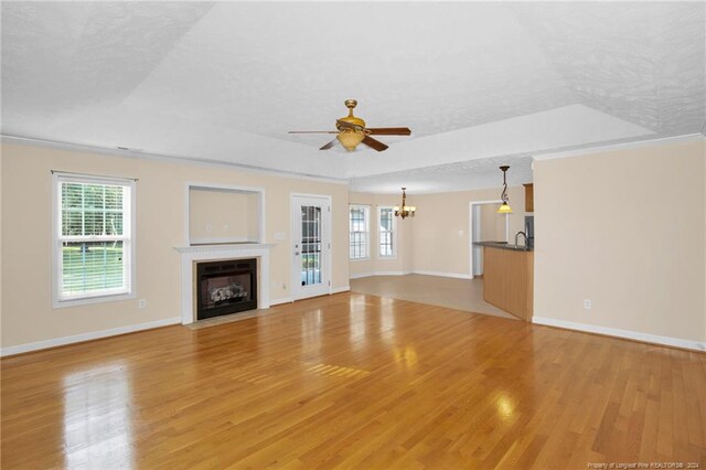 unfurnished living room featuring a raised ceiling, a wealth of natural light, ceiling fan with notable chandelier, and light wood-type flooring