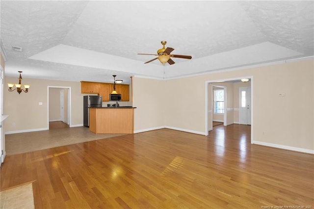 unfurnished living room with ceiling fan with notable chandelier, a textured ceiling, a tray ceiling, and light hardwood / wood-style flooring