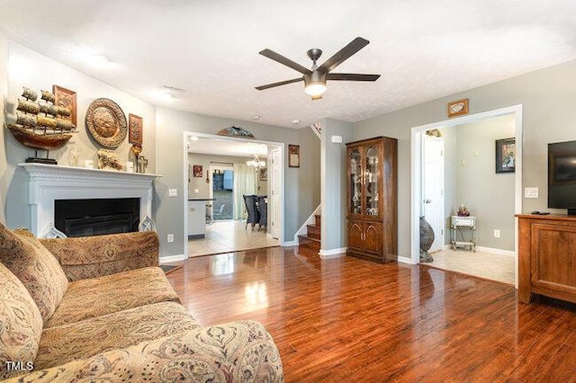 living room featuring stairway, a glass covered fireplace, wood finished floors, baseboards, and ceiling fan with notable chandelier