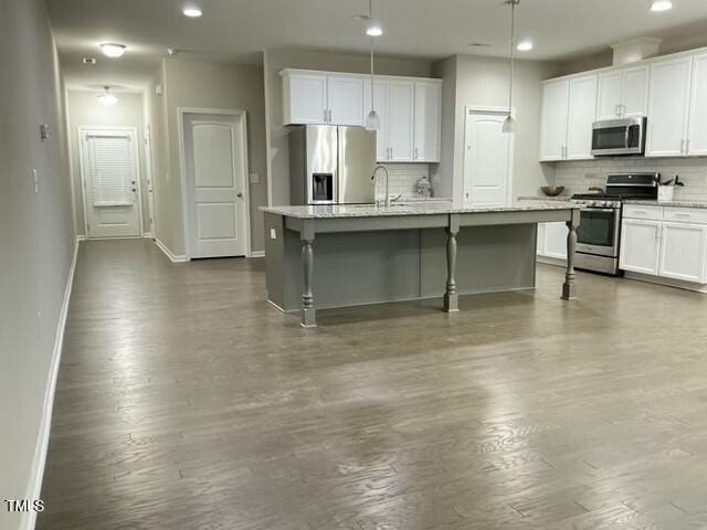 kitchen featuring appliances with stainless steel finishes, an island with sink, white cabinets, hanging light fixtures, and light stone countertops