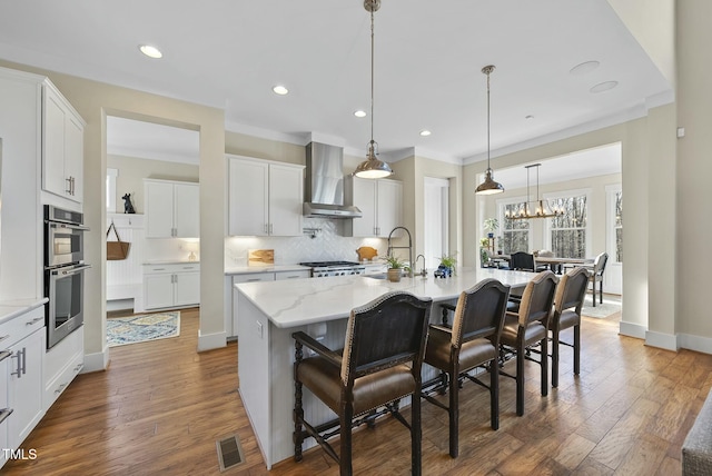 kitchen featuring wall chimney exhaust hood, a kitchen island with sink, sink, white cabinets, and a breakfast bar area