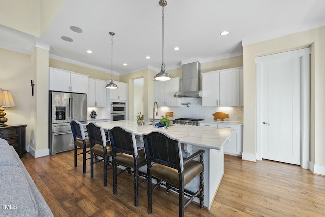 kitchen with white cabinets, wall chimney range hood, a kitchen island with sink, and appliances with stainless steel finishes