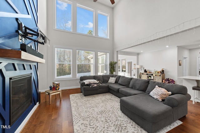 living area with dark wood-style flooring, a glass covered fireplace, a towering ceiling, and baseboards