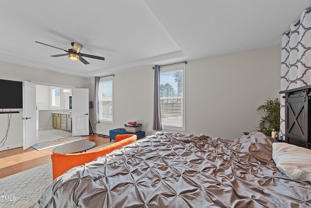 bedroom with ceiling fan, wood finished floors, baseboards, a tray ceiling, and ensuite bath