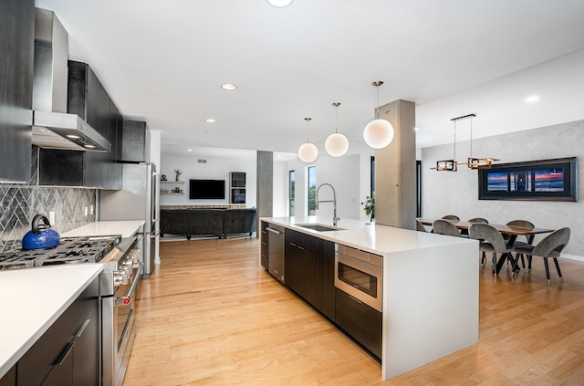 kitchen with pendant lighting, a kitchen island with sink, wall chimney range hood, sink, and appliances with stainless steel finishes