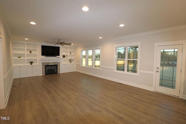 unfurnished living room with dark wood-type flooring, a stone fireplace, ceiling fan, built in features, and ornamental molding