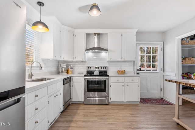 kitchen with sink, stainless steel appliances, wall chimney range hood, decorative light fixtures, and white cabinets