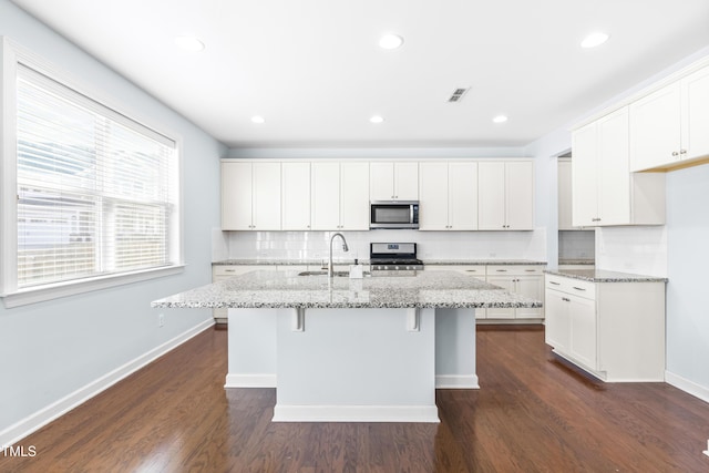 kitchen with light stone countertops, decorative backsplash, stainless steel appliances, white cabinetry, and an island with sink