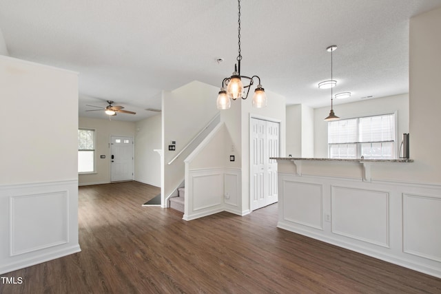 unfurnished living room with a decorative wall, a ceiling fan, dark wood-type flooring, wainscoting, and stairway