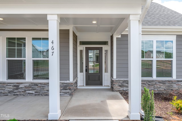 view of exterior entry with stone siding, roof with shingles, and a porch