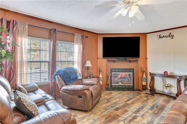 living area featuring ceiling fan, a textured ceiling, a fireplace with flush hearth, wood finished floors, and ornamental molding