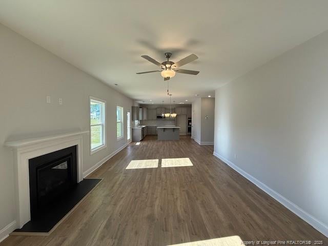 unfurnished living room featuring a ceiling fan, baseboards, dark wood-style flooring, and a glass covered fireplace