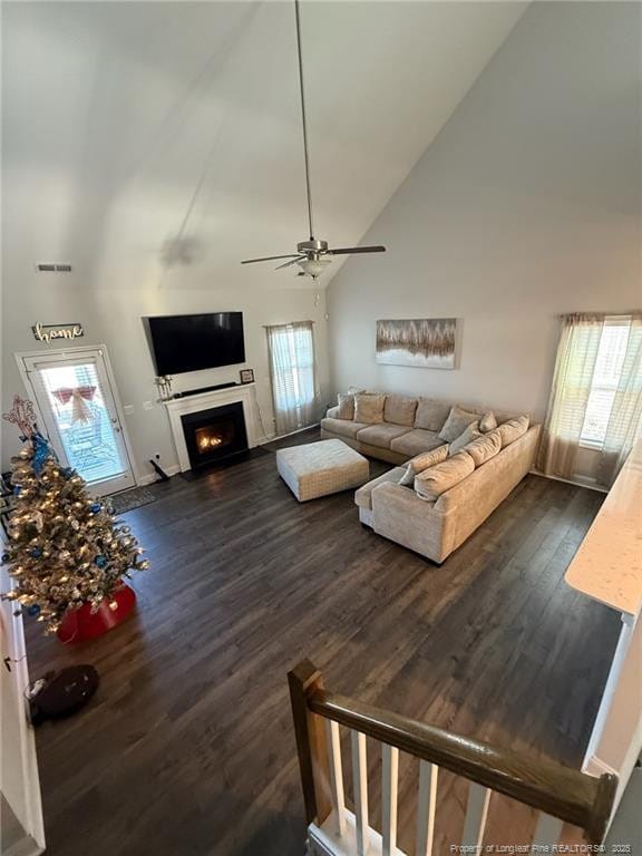 living area featuring high vaulted ceiling, dark wood finished floors, a lit fireplace, and a ceiling fan