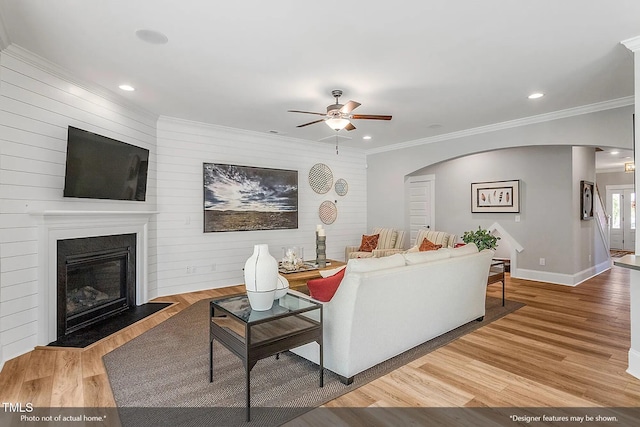 living room with light wood finished floors, a fireplace with flush hearth, ornamental molding, arched walkways, and a ceiling fan