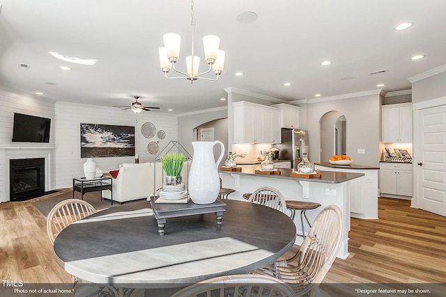 dining area with light wood finished floors, crown molding, ceiling fan with notable chandelier, arched walkways, and a glass covered fireplace