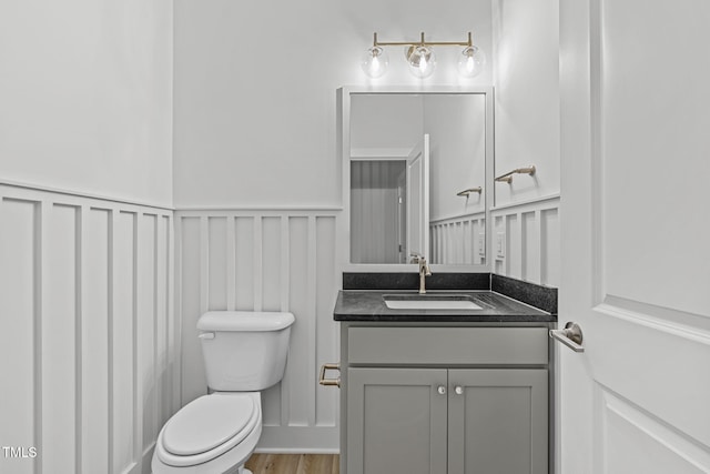 half bathroom with a decorative wall, toilet, wainscoting, vanity, and wood finished floors