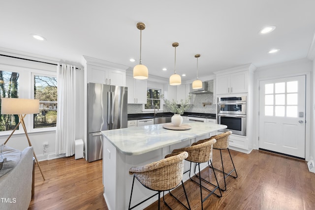 kitchen featuring white cabinets, appliances with stainless steel finishes, wall chimney exhaust hood, hanging light fixtures, and crown molding