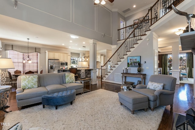 living room featuring dark wood-type flooring, ceiling fan with notable chandelier, crown molding, and ornate columns