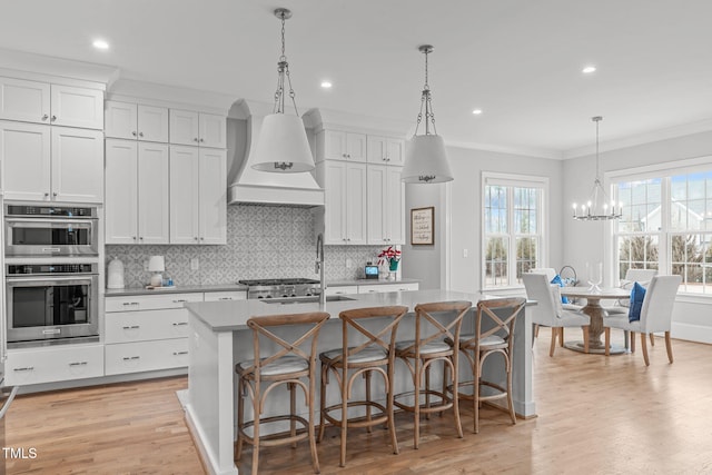 kitchen featuring a center island with sink, hanging light fixtures, custom range hood, double oven, and white cabinets