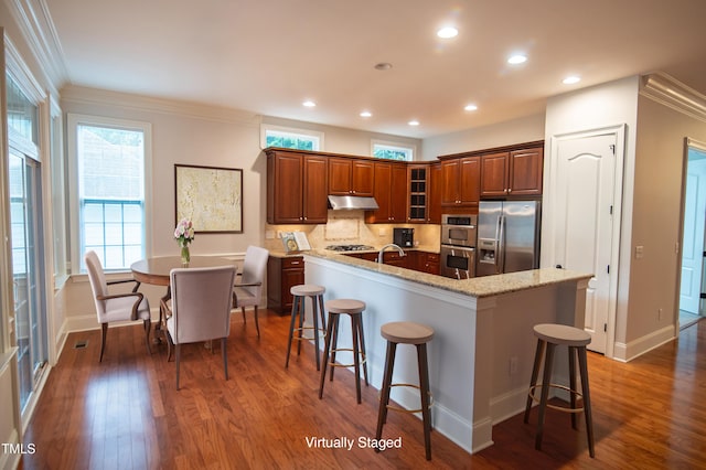 kitchen featuring a breakfast bar, a center island with sink, under cabinet range hood, appliances with stainless steel finishes, and dark wood-style flooring