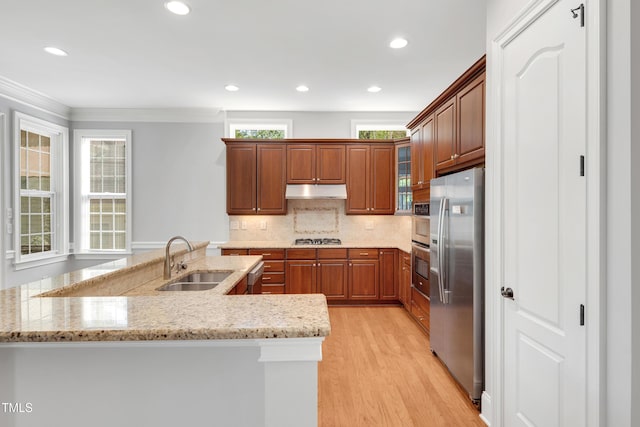 kitchen featuring a sink, light wood-style floors, under cabinet range hood, brown cabinets, and stainless steel appliances