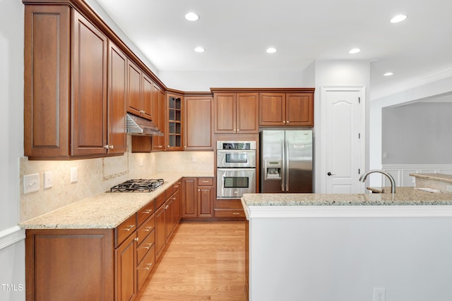 kitchen with glass insert cabinets, brown cabinetry, appliances with stainless steel finishes, light stone counters, and under cabinet range hood