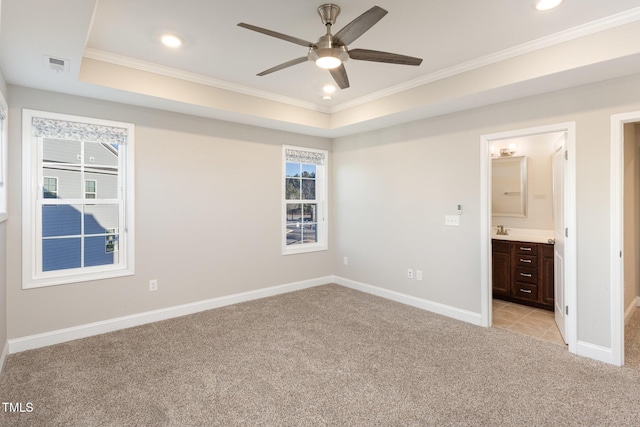 unfurnished bedroom featuring a raised ceiling, ensuite bath, light colored carpet, crown molding, and ceiling fan